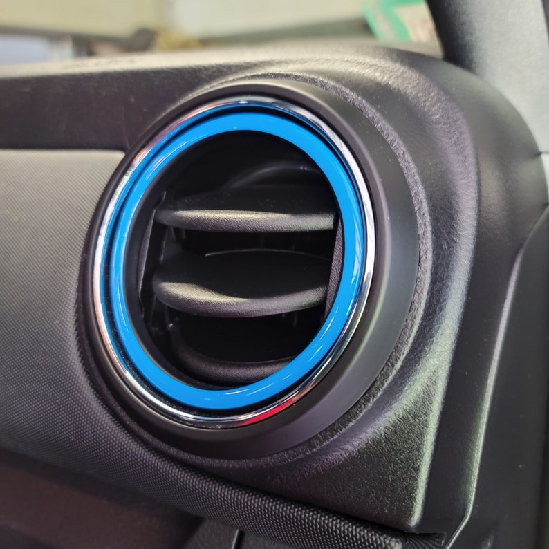 May include: A close-up of a car's air vent with a blue and silver trim ring. The vent is made of black plastic and has a series of horizontal slats.