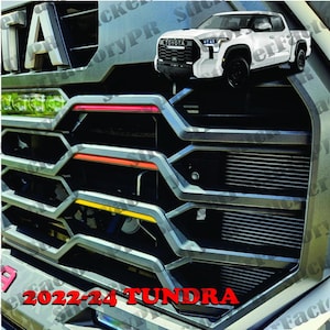 Puede incluir: Primer plano de una parrilla de camión gris con una franja de acento roja, naranja y amarilla. La parrilla tiene un patrón de panal y el texto "2022-24 TUNDRA" es visible en la parte inferior. Un camión de recogida Toyota Tundra blanco es visible en el fondo.