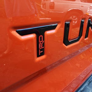 以下が含まれることがあります： オレンジ色の車の黒と赤の「TRD」エンブレムのクローズアップ。エンブレムは車の後ろにあり、「TUNDRA」の文字の一部です。