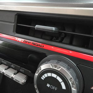 May include: Close-up of a car's dashboard with a black and silver knob labeled "OFF". The dashboard has a red accent stripe and the text "TRD OFF ROAD" in white letters.