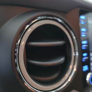 May include: Close-up of a car's air vent with a silver ring around the opening. The vent is black and has a series of horizontal slats.
