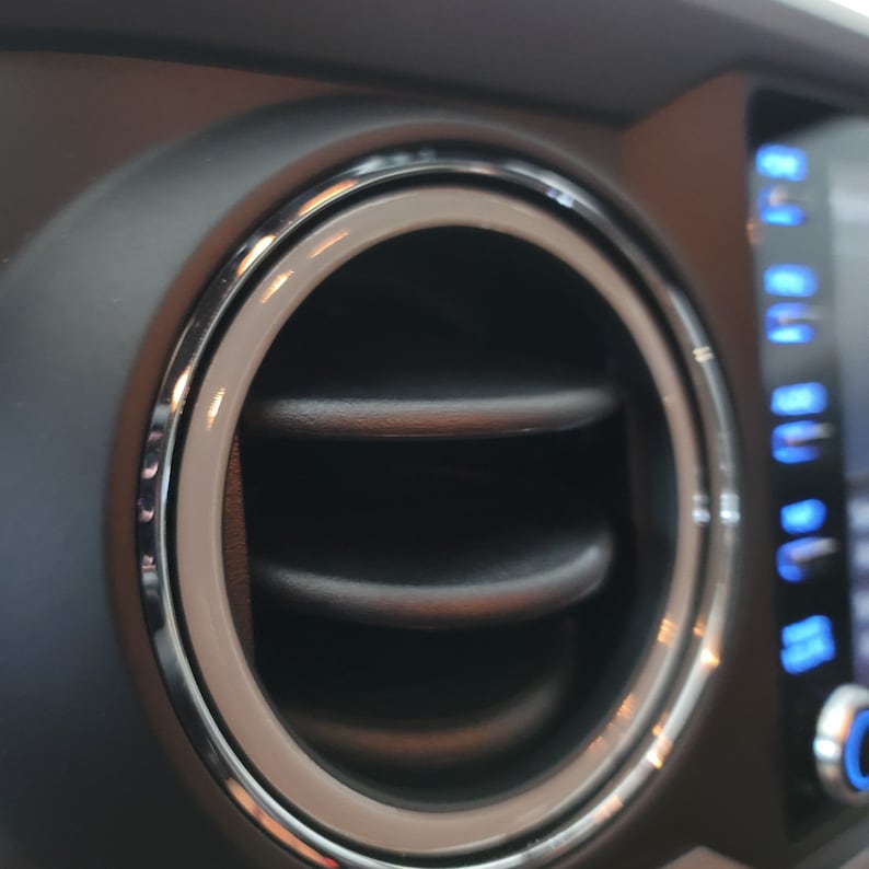 May include: Close-up of a car's air vent with a silver ring around the opening. The vent is black and has a series of horizontal slats.