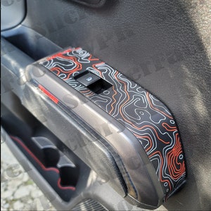 May include: A black car door panel with a red and white topographic map design. The panel has a window control switch and a cup holder.