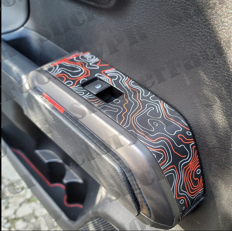 May include: A black car door panel with a red and white topographic map design. The panel has a window control switch and a cup holder.