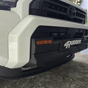 Puede incluir: Primer plano de la parte delantera de un Toyota 4Runner blanco. La parrilla negra presenta el emblema de Toyota. El parachoques es negro con el texto "4Runner" en blanco. Luces ámbar visibles.