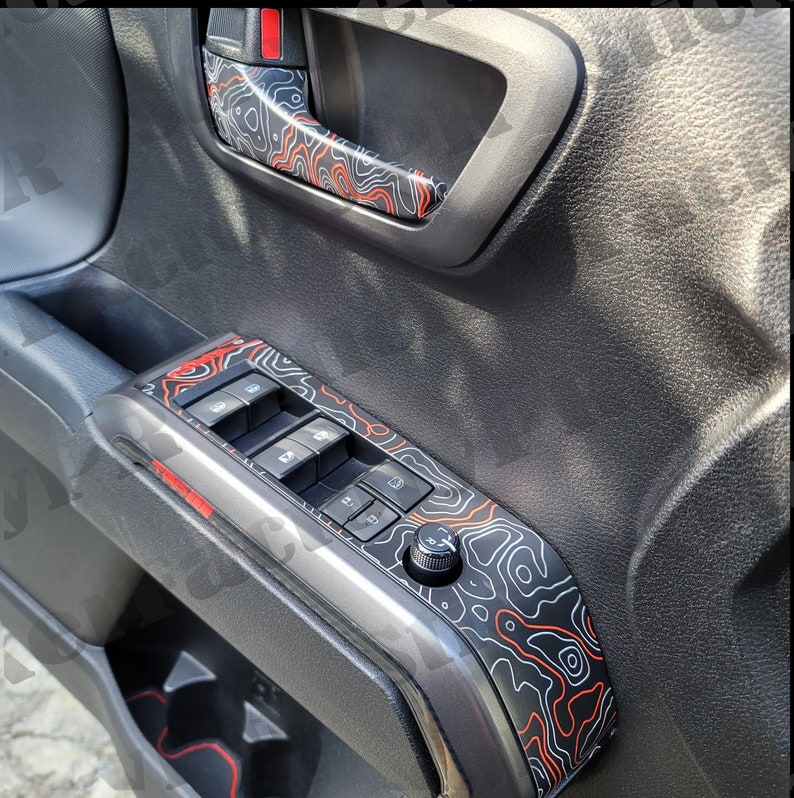 May include: A black and gray car door panel with a red and black topographic pattern. The panel features a window control panel with four buttons and a knob.