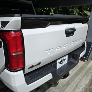 Puede incluir: Camioneta Toyota Tacoma blanca con una puerta trasera negra y un emblema 4x4 negro. La camioneta tiene un marco de matrícula negro con las letras "SF" en un círculo blanco.