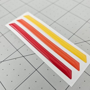 K&ouml;nnte beinhalten: Drei Vinylstreifen in Rot, Orange und Gelb auf wei&szlig;em Untergrund. Die Streifen sind gl&auml;nzend und haben eine erhabene Textur.
