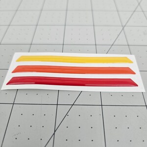 K&ouml;nnte beinhalten: Drei Vinylstreifen in den Farben Rot, Orange und Gelb. Die Streifen befinden sich auf einem wei&szlig;en Hintergrund und sind bereit, auf ein Auto oder eine andere Oberfl&auml;che aufgebracht zu werden.