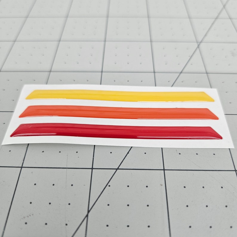 K&ouml;nnte beinhalten: Drei Vinylstreifen in den Farben Rot, Orange und Gelb. Die Streifen befinden sich auf einem wei&szlig;en Hintergrund und sind bereit, auf ein Auto oder eine andere Oberfl&auml;che aufgebracht zu werden.