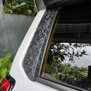 May include: A white vehicle exterior pillar trim piece featuring a dark grey and black forged carbon fibre pattern. The trim is positioned vertically along the edge of the rear window, which displays horizontal defroster lines and Toyota branding.