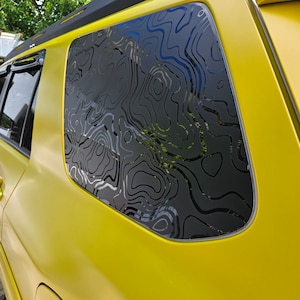 May include: A bright yellow vehicle features a rear side window adorned with a black topographic map decal. The decal consists of intricate, flowing contour lines that cover the entire glass surface, contrasting against the vibrant yellow paint.