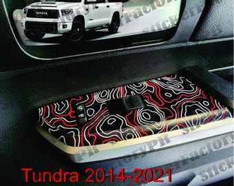 May include: A black interior trim overlay for a Toyota Tundra 2014-2021 featuring a topographic map pattern with white and red contour lines. The decal is applied to the center console area, which includes a black lock button and a rectangular switch.