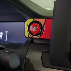May include: A red engine start stop button with a black ring and the word "TRD" in white letters. The button is surrounded by a yellow, orange, and red stripe design.
