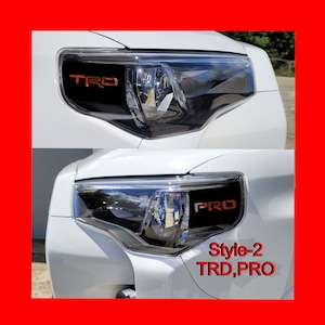 以下が含まれることがあります： 白い車両用ヘッドライトアセンブリ。上部にオレンジ色の「TRD」、下部にオレンジ色の「PRO」の文字が入った黒いインサートが特徴です。画像右下には「Style-2 TRD, PRO」という赤い文字が印刷されています。