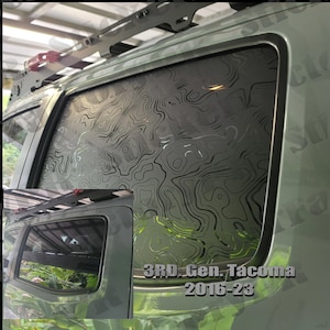 May include: A close-up of a green truck with a black window tint. The window tint has a topographic map design. The truck has a roof rack and the text "3RD Gen. Tacoma 2016-23" is visible on the side of the truck.