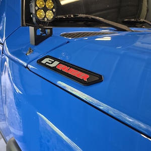 以下が含まれることがあります： 鮮やかな青色の車両のボンネットに、黒と赤のFJ Cruiserエンブレムが取り付けられています。エンブレムには白でFJ、赤でCRUISERという文字が記されています。ボンネットの角には、4つの黄色い円形レンズを備えた黒い補助ライトポッドが装着されています。