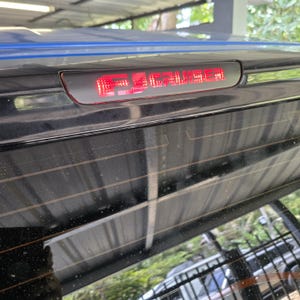 Peut inclure: Un boîtier de troisième feu stop automobile noir avec un affichage LED rouge illuminé indiquant FJ CRUISER. Le feu est intégré dans la partie supérieure de la lunette arrière d'un véhicule, qui présente des lignes de dégivrage horizontales.