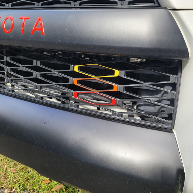Pu&ograve; includere: Una griglia per veicoli a nido d'ape nera con il testo rosso TOYOTA nella parte superiore. Tre inserti esagonali impilati in giallo, arancione e rosso sono posizionati al centro della struttura a griglia in plastica nera su un veicolo bianco.