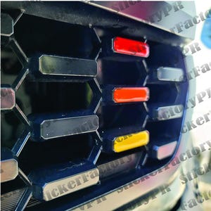 Könnte beinhalten: Nahaufnahme eines schwarzen Wabengrills mit drei roten, gelben und schwarzen Akzentstreifen. Die Streifen sind horizontal über dem Grill angeordnet.