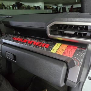 May include: A black and silver dashboard trim piece with the word "4RUNNER" in red. The trim features a topographic map design in black, red, yellow, and orange. The trim is installed on a black dashboard.