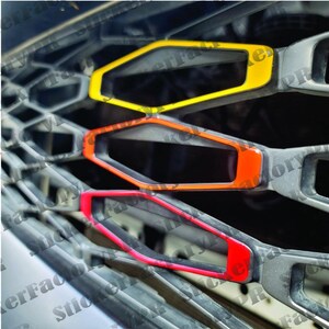 Pu&ograve; includere: Un set di inserti per griglia auto con tre accenti esagonali in giallo, arancione e rosso. Questi rivestimenti colorati sono fissati sulle singole aperture di una struttura della griglia anteriore del veicolo grigio scuro con motivo a rombi.