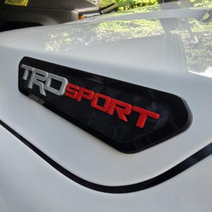 Könnte beinhalten: Eine weiße Fahrzeugmotorhaube mit einem schwarzen, eingelassenen Lufthutzen-Einsatz. Der Einsatz zeigt den erhabenen, dreidimensionalen Schriftzug TRD in Silber und SPORT in Rot mit einer glänzenden Oberfläche, die sich von der weißen Metalloberfläche abhebt.