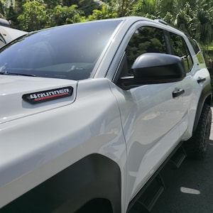 May include: A white SUV with black accents, including the side mirrors and fender flares. The vehicle has the text "4Runner" in red and black on the front. The tires are black and the vehicle is parked outside.