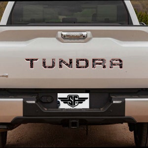 Puede incluir: La vista trasera de una camioneta plateada con la palabra TUNDRA en letras grandes y negritas en el portón trasero. Las letras tienen un patrón de mapa topográfico en blanco y negro con un borde rojo fino. Un emblema 4x4 está a la izquierda.