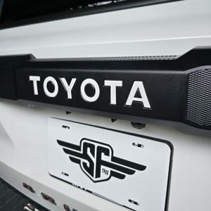 May include: A white vehicle tailgate featuring a prominent black trim piece with the word TOYOTA in bold white capital letters. Below the trim, a white number plate displays a black graphic of a shield with wings and the text SF 98.