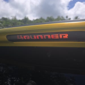 May include: A yellow vehicle exterior component featuring a horizontal black rectangular light housing. The light displays the illuminated red text 4RUNNER in a dot-matrix style, set against a bright yellow body panel under a cloudy sky.