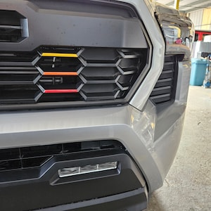 K&ouml;nnte beinhalten: Nahaufnahme des K&uuml;hlergrills eines silbernen Pickups mit einem schwarzen Wabenmuster und drei horizontalen Streifen in Gelb, Orange und Rot.