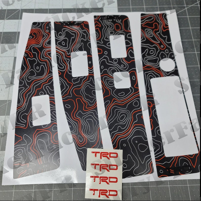 May include: Set of four black and white car interior trim decals with a red topographic pattern. The decals are designed to fit around the window switches and door handles. The set also includes four red "TRO" decals.