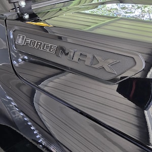 Puede incluir: Un guardabarros de vehículo en color negro brillante con un emblema negro mate que presenta el texto iFORCE MAX en letras en relieve. El emblema está situado en un panel empotrado en el lateral de la carrocería, mostrando un acabado oscuro y elegante.