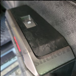 May include: Close-up of a black car door panel with a window control switch and a textured surface. The panel has a silver trim and a red accent.