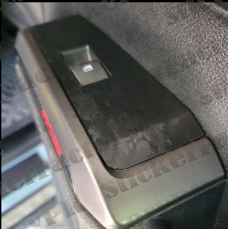 May include: Close-up of a black car door panel with a window control switch and a textured surface. The panel has a silver trim and a red accent.