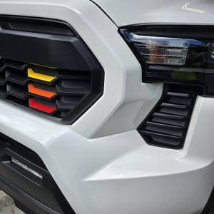 Könnte beinhalten: Nahaufnahme eines weißen Pick-up-Trucks mit einem schwarzen Kühlergrill und einem schwarzen und gelben Akzentstreifen. Der Kühlergrill hat ein Wabenmuster und der Akzentstreifen besteht aus vier dünnen Streifen.