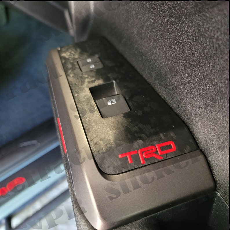 May include: A black car interior panel with a carbon fiber pattern and a red "TRD" logo. The panel has a window switch and two other buttons.