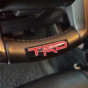 May include: Close-up of a black leather steering wheel with a red "TRD" logo.