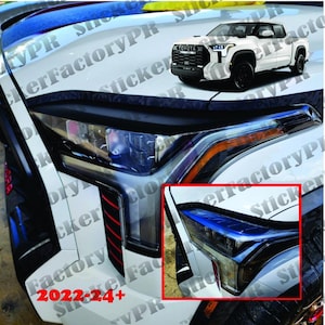 May include: Close-up of a white pickup truck's front headlight with a black and blue accent. The truck has a black grill with the word "TOYOTA" in white letters. The truck is parked on a black surface.