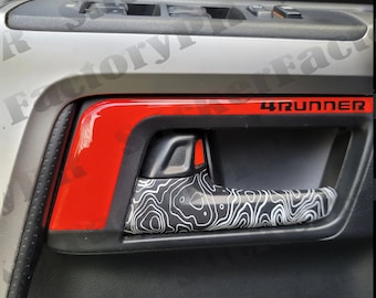 Può includere: Un gruppo maniglia interna per portiera di veicolo con una finitura rossa lucida che riporta la scritta 4RUNNER in nero. La maniglia stessa è decorata con un motivo a mappa topografica in bianco e nero su un pannello porta grigio scuro.