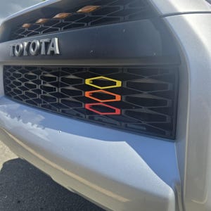 Op de afbeelding: Een zilverkleurige Toyota-voertuiggrille met een zwart honingraatpatroon en een gestapeld embleem bestaande uit drie zeshoekige contouren in geel, oranje en rood, horizontaal geplaatst aan de rechterkant van de grille.