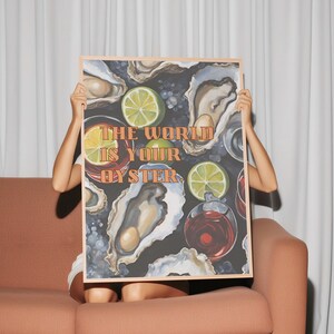 May include: A framed art print featuring an oyster-themed illustration with the text "THE WORLD IS YOUR OYSTER." The artwork includes images of oysters, lime slices, and drinks, set against a dark background. The frame is a light wood color.