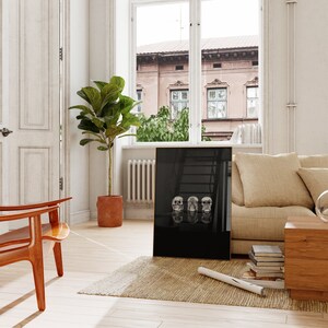 May include: A black framed art print displaying three white skull sculptures. The skulls are in a row, each with a different expression. The print is propped against a wall in a room with a window and a potted plant.