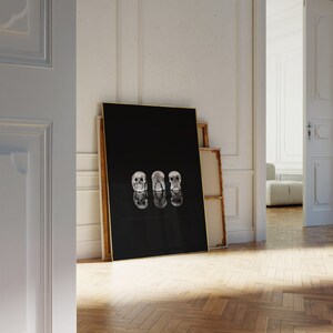 May include: A black framed art print with three skull illustrations, each with a different hand gesture. The gold-framed print is leaning against a white wall. The print is approximately 100 cm in height.