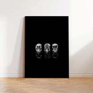 May include: A black canvas print showing three silver skulls in the 'see no evil, hear no evil, speak no evil' pose. The skulls are reflected on the black surface. The artwork is displayed against a white wall and wooden floor.
