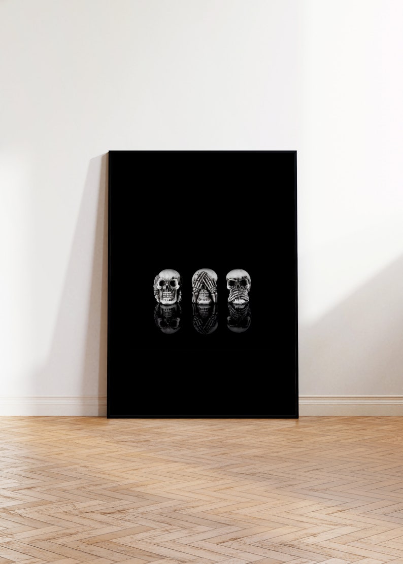 May include: A black canvas print showing three silver skulls in the 'see no evil, hear no evil, speak no evil' pose. The skulls are reflected on the black surface. The artwork is displayed against a white wall and wooden floor.