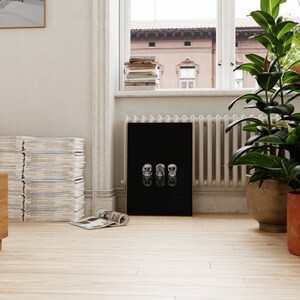 May include: A black framed art print with three skull designs. The artwork is displayed on a light wood floor. A stack of magazines and a potted plant are in the background. The room has a window and a radiator.