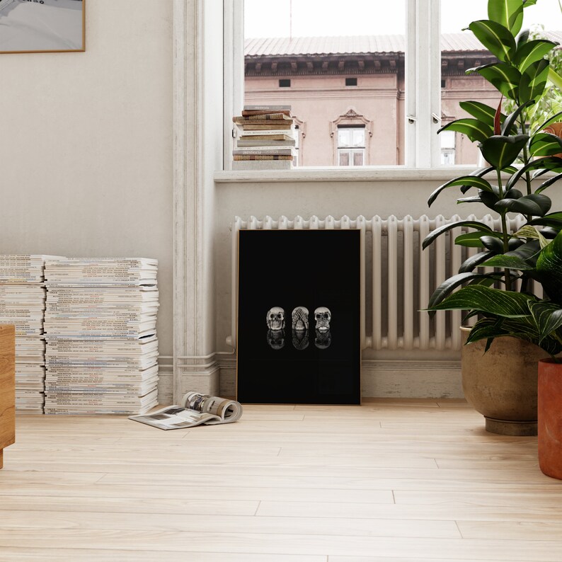 May include: A black framed art print with three skull designs. The artwork is displayed on a light wood floor. A stack of magazines and a potted plant are in the background. The room has a window and a radiator.