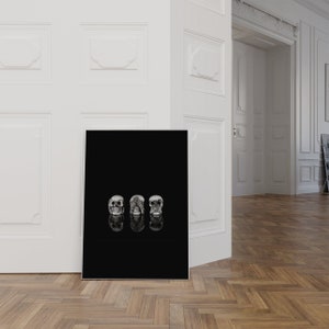 May include: A monochrome art print showing three skull sculptures. Each skull embodies a different concept: see no evil, hear no evil, and speak no evil. The skulls are reflected on a black surface.